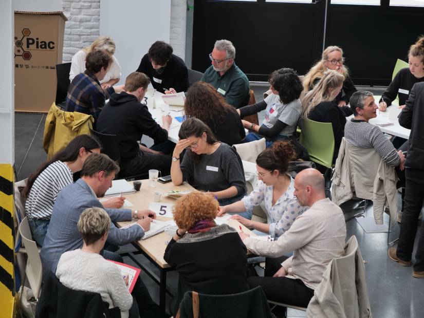 Plusieurs groupes de personnes attablées se concentrent sur un travail écrit, d'autres discutent dans une ambiance décontractée